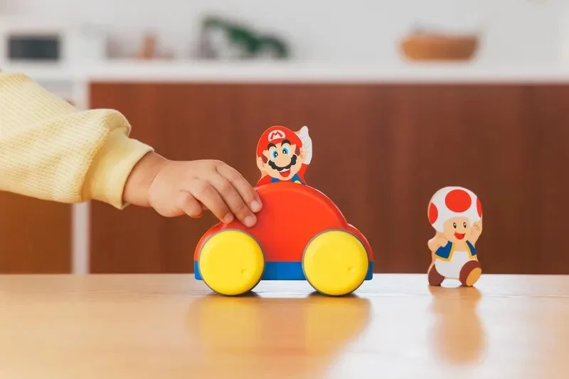 Ein Kind spielt mit einem roten Mario-Auto und einer Toad-Figur auf einem Tisch.